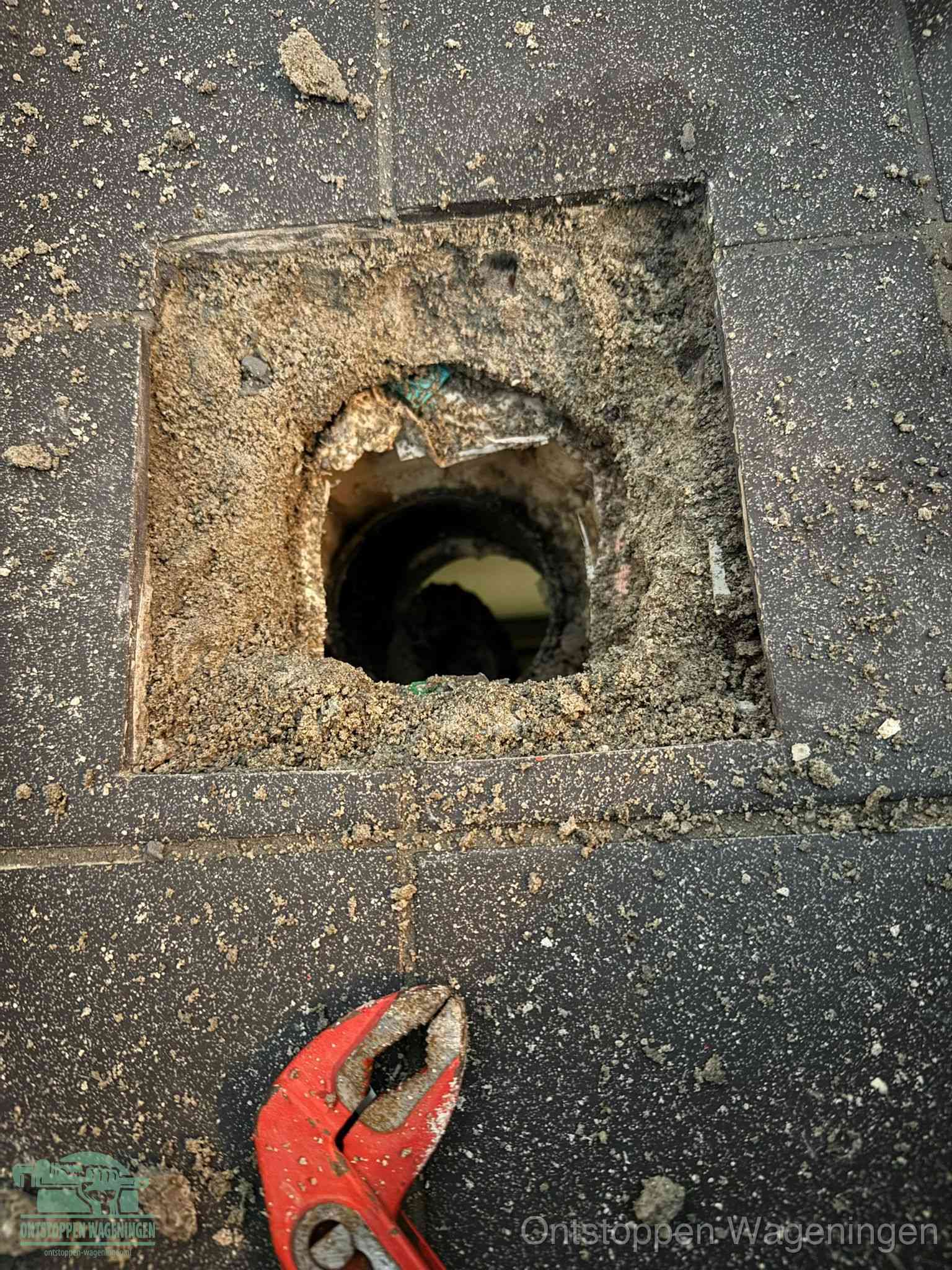 Een geopend rioolputje in een betontegelvloer met losgemaakt rooster en een zichtbare ronde afvoerbuis, het achterhalen waar de afvoerverstopping zit in Wageningen