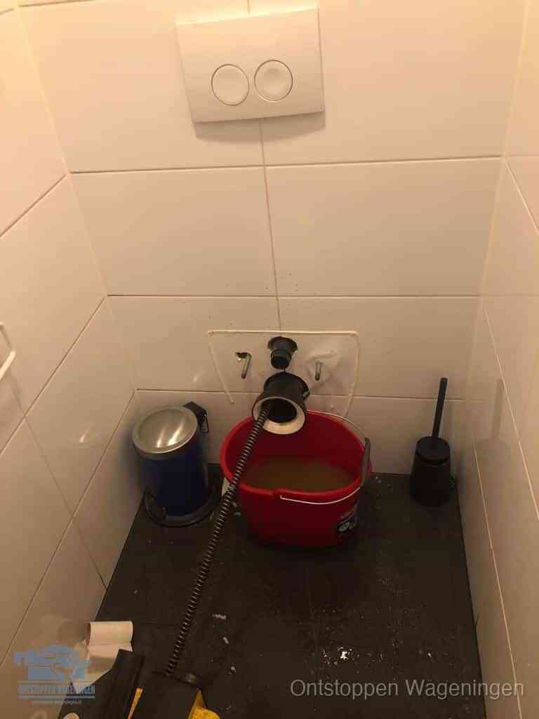 Een ontstopte wand-wc met verwijderde toiletpot waar een ontstoppingsveer in de wanduitgang verdwijnt, het achterhalen waar de verstopping zit in Wageningen
