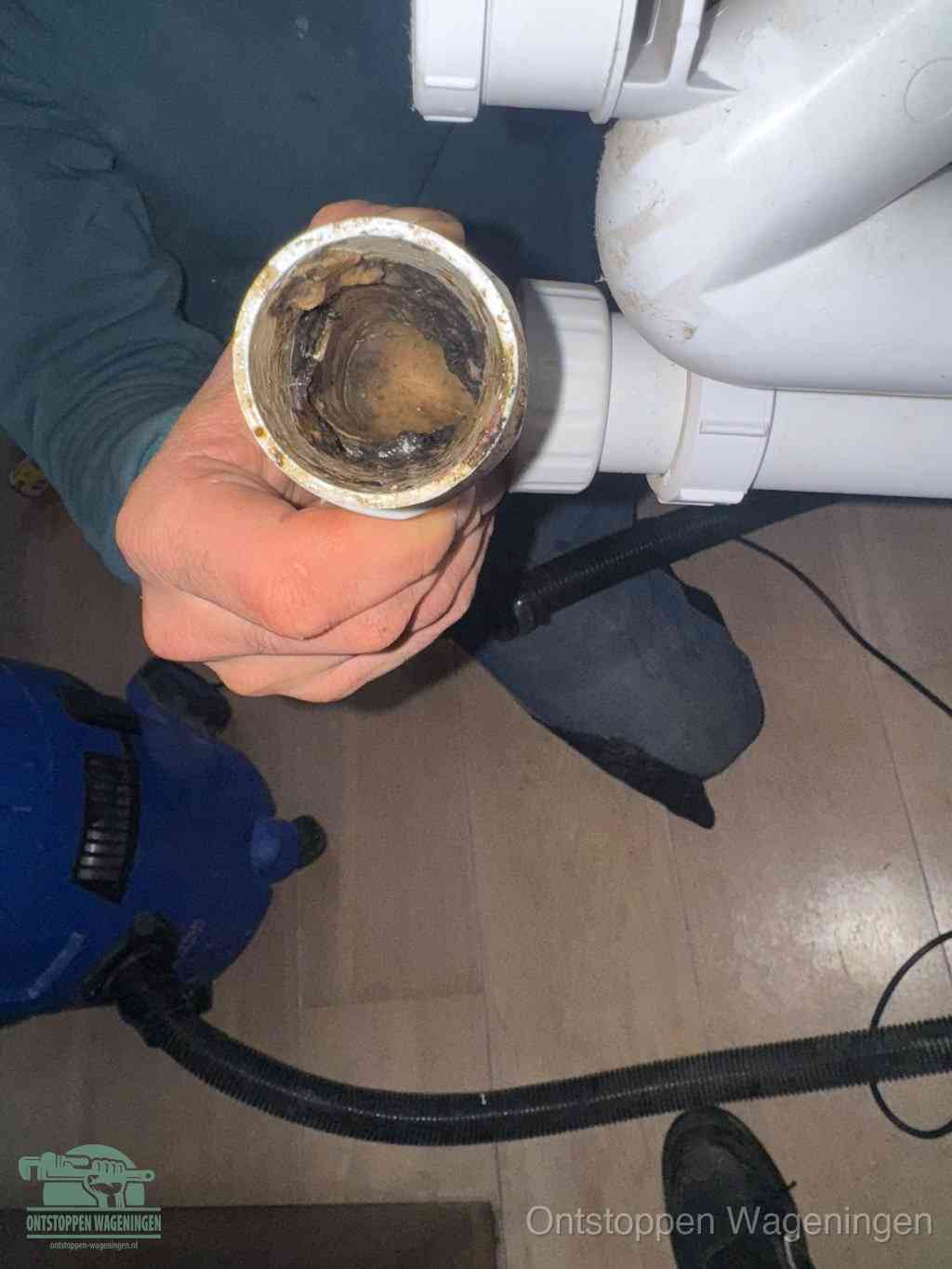 Een hand houdt een losgekoppeld PVC-afvoerbuisstuk vast dat van binnen volledig is dichtgeslibd met vet, vuil en aanslag, de meest voorkomende oorzaak van een verstopte afvoer in Wageningen