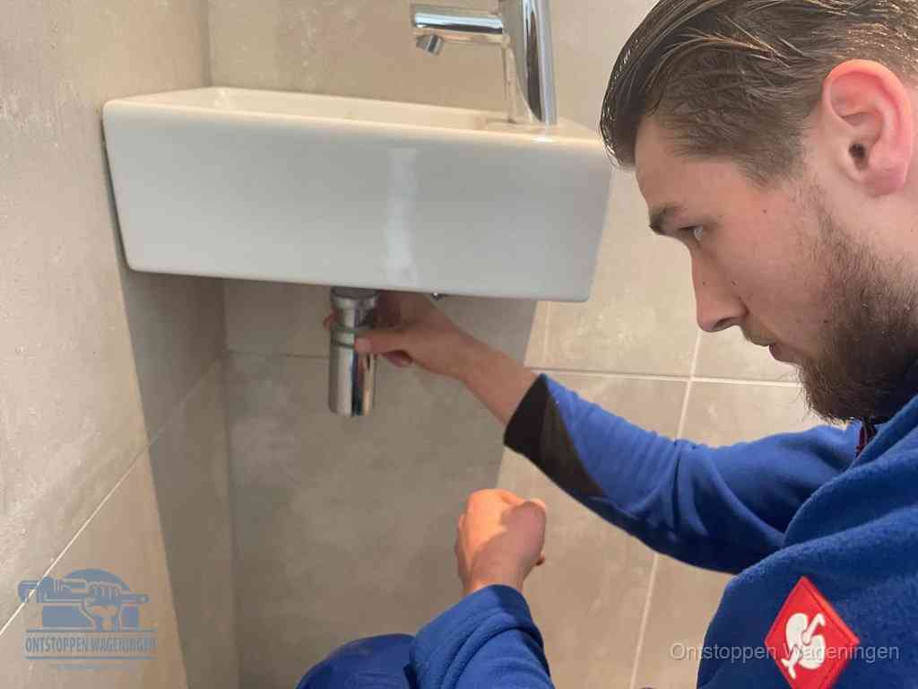 Een monteur draait met de hand de zwanenhalssifon onder een fonteinwasbak in de toiletruimte los om de bron van rioollucht in Wageningen te vinden