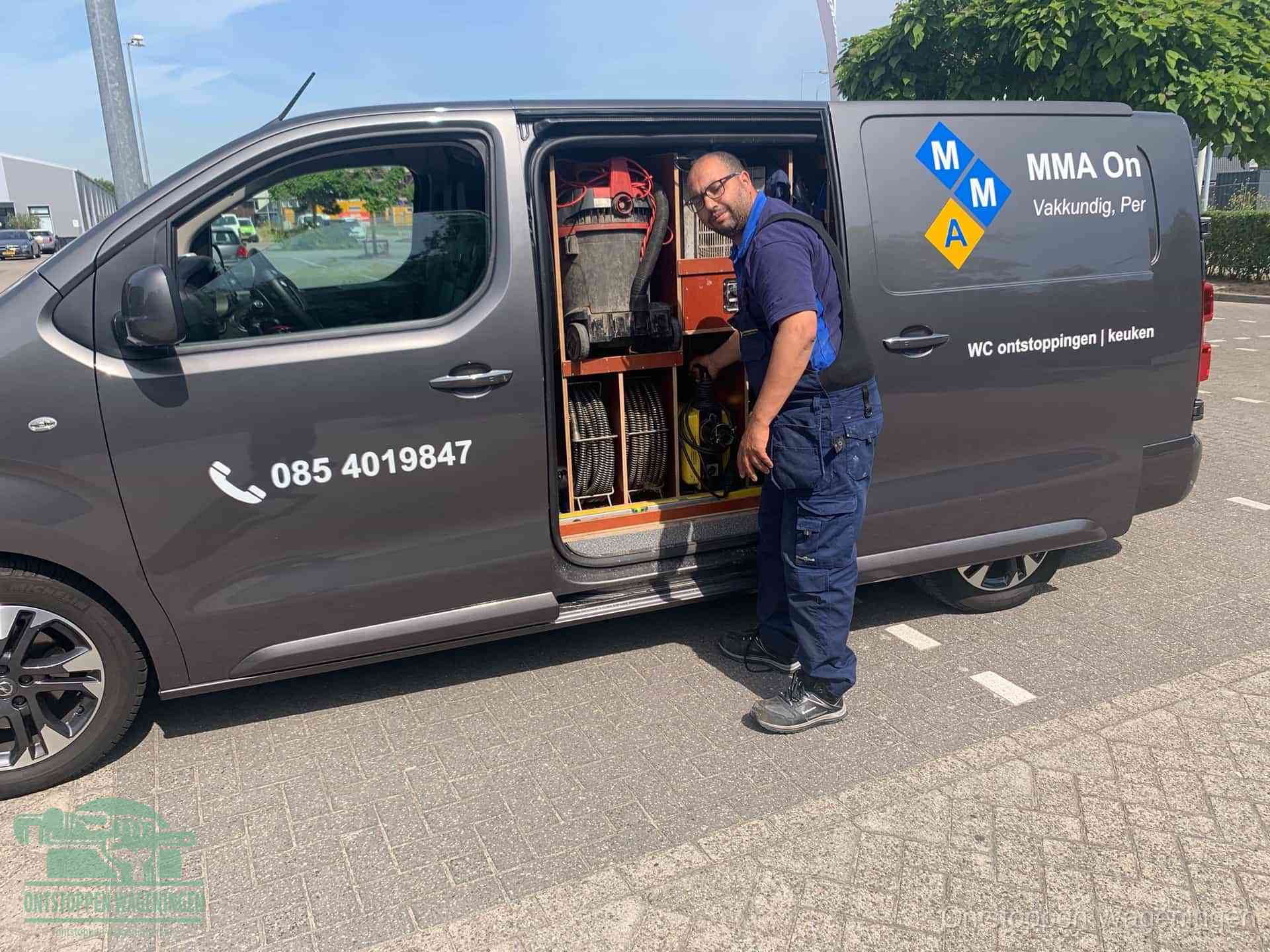 Een monteur bij de open schuifdeur van zijn bedrijfsbus op een parkeerplaats, klaar om de oorzaak van terugkerende afvoerverstoppingen aan te pakken in Wageningen