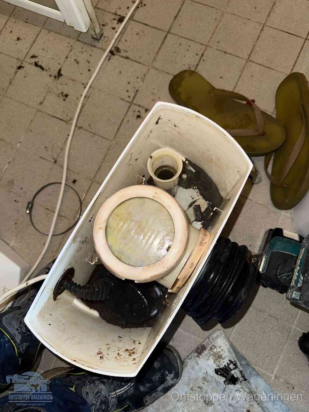 Een gedemonteerde toiletpot op de vloer met ontstoppingsveer en gereedschap ernaast, het verwijderen van een vastzittend voorwerp uit de wc in Wageningen