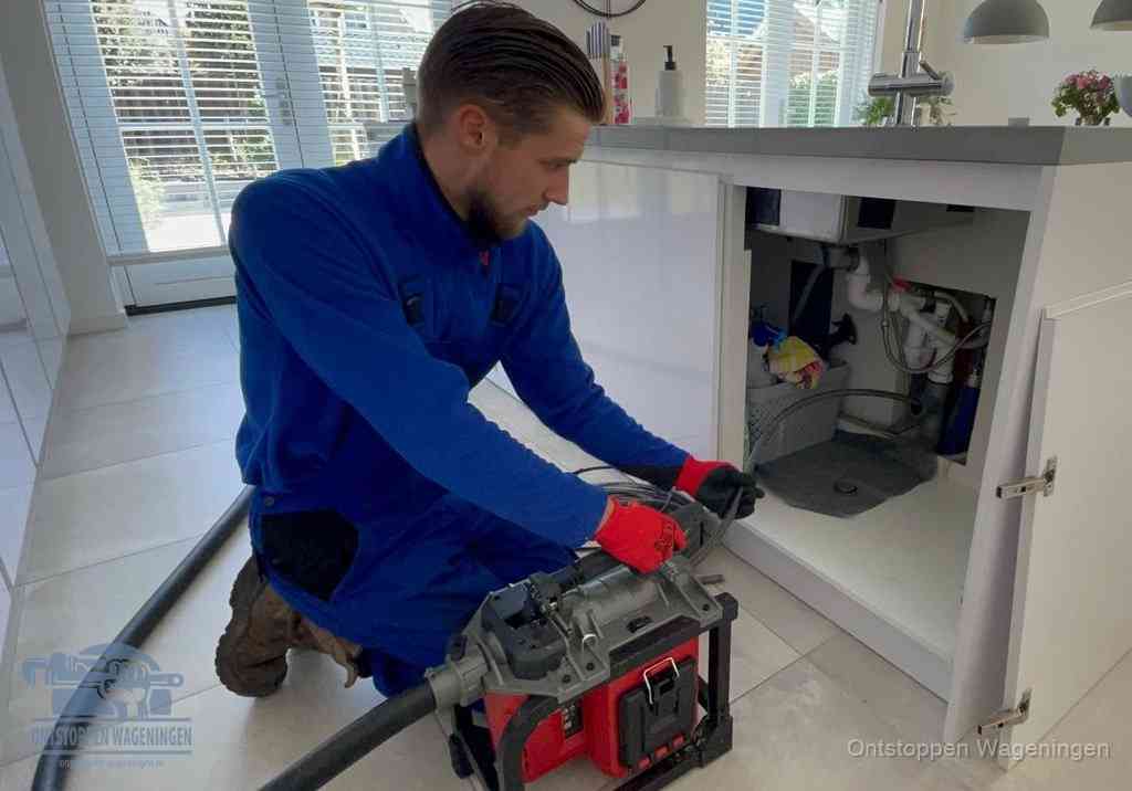 Een monteur voert een professionele elektrische ontstoppingsmachine in de afvoer onder de spoelbak tijdens het gootsteen ontstoppen in Wageningen